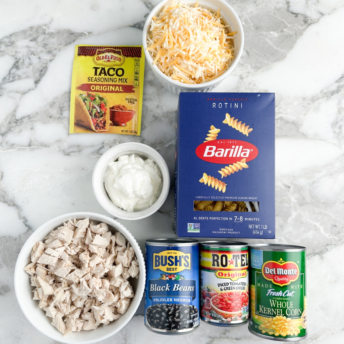 Rotini pasta, taco seasonings, bowl of shredded cheese, diced chicken, sour cream, cans of beans, tomatoes, and corn.