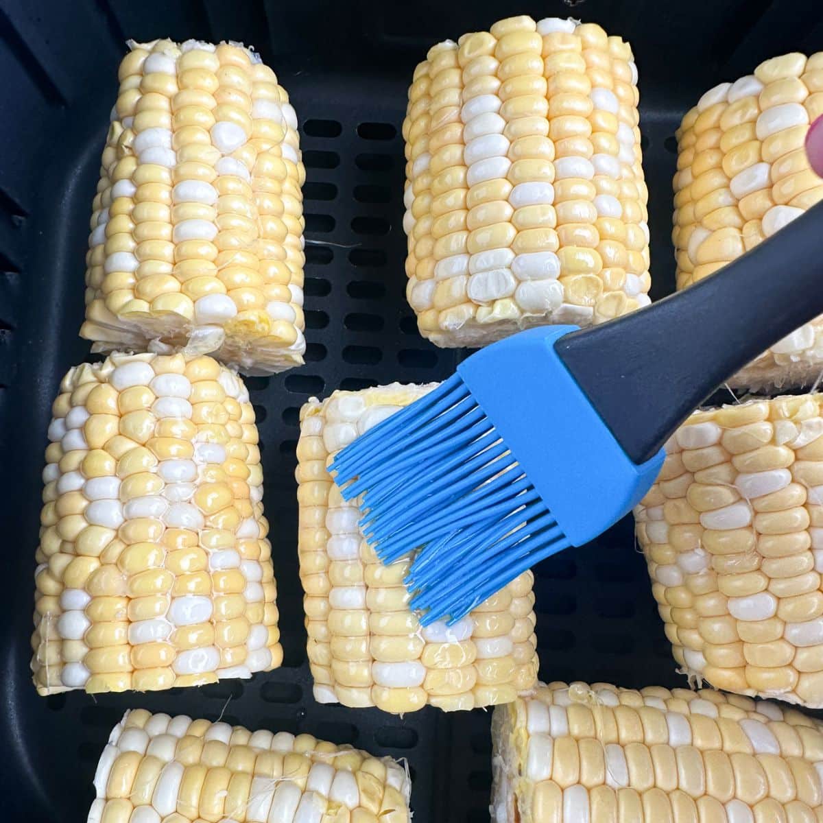 Corn in air fryer basket with oil being brushed on. 