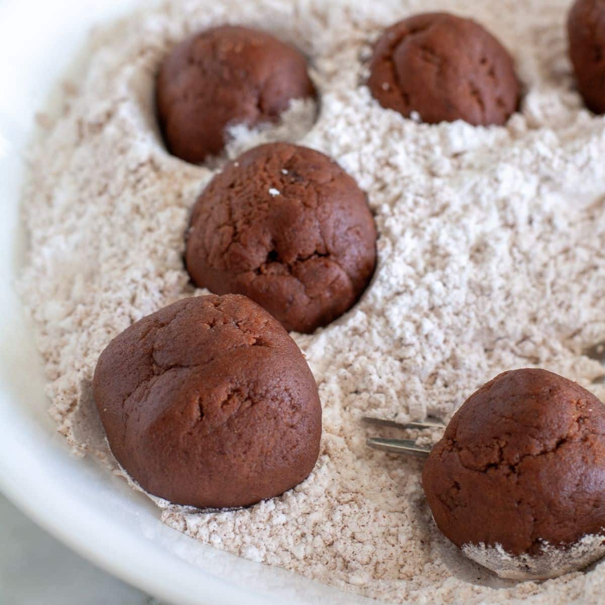 Chocolate cookie dough balls in a bowl of powdered sugar.