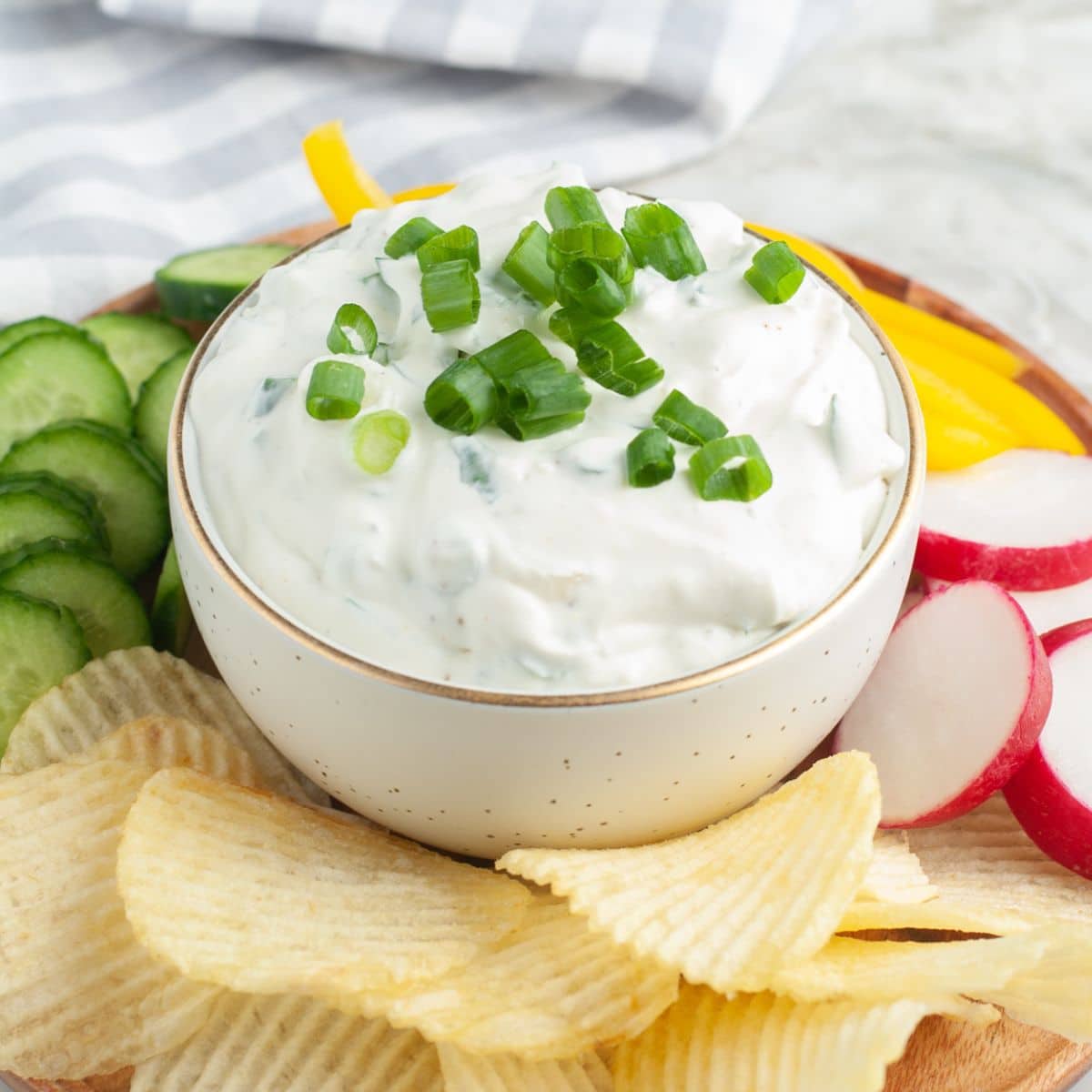 Bowl of green onion dip with potato chips, radish, and sliced cucumbers.