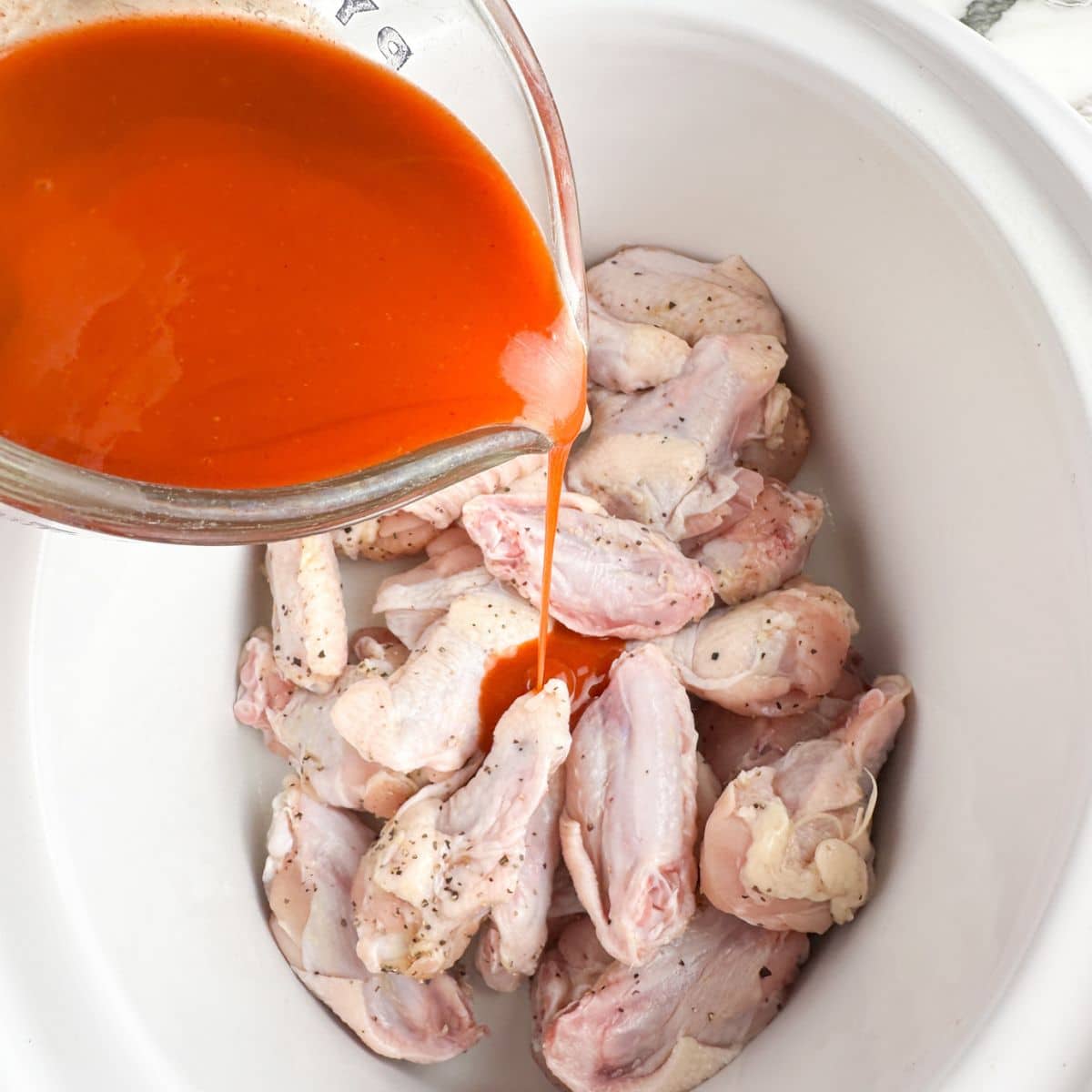 Slow cooker full of chicken wings with buffalo sauce being poured on them. 