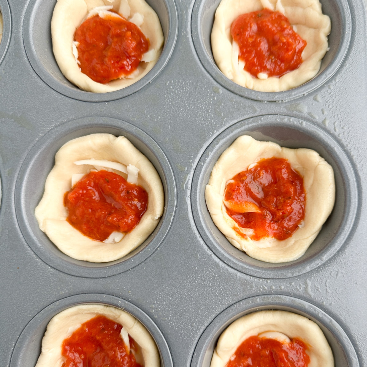 Muffin tin with pizza dough, cheese, and pizza sauce. 